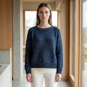 May include: A woman wearing a dark blue crew neck sweater with a textured knit. The sweater is paired with white pants. The background features wooden paneling and large windows, creating a bright, natural setting.