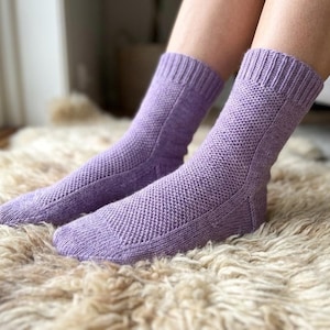 Könnte beinhalten: Ein Paar lavendelfarbene Strickstrümpfe mit einem strukturierten Muster. Die Socken haben gerippte Bündchen und werden auf einem flauschigen, cremefarbenen Teppich präsentiert. Die Socken sind auf Komfort und Wärme ausgelegt.