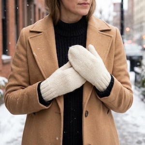 Può includere: Moffole in maglia color crema con polsini neri, indossate da una persona con un cappotto color cammello e un maglione nero. La scena è all'aperto con neve che cade e un edificio in mattoni sullo sfondo.