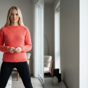 May include: A coral-colored crochet sweater with a crew neck and long sleeves. The sweater is paired with black leggings. The image is taken indoors with natural light.