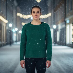 May include: A person wearing a green jumper and black trousers stands on a snow-covered street. The jumper has a ribbed hem and cuffs. Snowflakes fall in front of a blurred background of buildings and streetlights.
