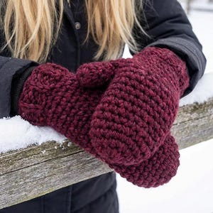 May include: A pair of burgundy crochet mittens. The mittens are thick and textured, with a visible stitch pattern. The mittens are resting on a wooden fence covered in snow. The background is blurred, suggesting an outdoor winter scene.