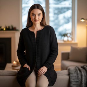 Può includere: Un cardigan lungo nero con tasche. Il cardigan è indossato sopra pantaloni colorati neutri. La persona è seduta su un divano grigio in un soggiorno con un camino e una finestra con vista sulla neve.