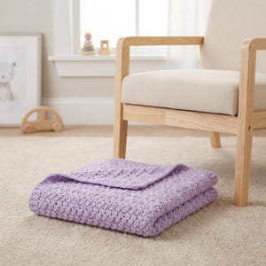 May include: A folded, light purple crochet blanket rests on a light beige carpet. The blanket is made of a textured yarn and is folded neatly. A wooden chair with a beige cushion is in the background.