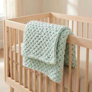 May include: A light green, chunky knit baby blanket draped over the side of a light wood crib. The blanket has a woven texture and a soft, plush appearance. The crib is empty, with a neutral-colored mattress.