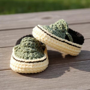 Op de afbeelding: Een paar handgemaakte gehaakte babyschoentjes. De schoenen hebben een groen bovenwerk, een crèmekleurige zool en zwarte accenten. De schoenen zijn gemaakt van zacht garen en lijken op miniatuur sneakers.