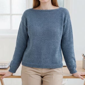 May include: A pale blue knitted jumper with a boat neck and long sleeves. The jumper is made of a textured knit fabric and has a ribbed hem and cuffs. The model is wearing beige trousers.