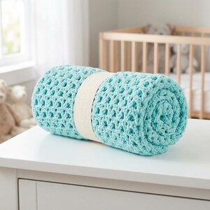 May include: A rolled-up, light blue crochet blanket secured with a cream-colored band. The blanket is made of a soft, open-weave material. The blanket is resting on a white surface.