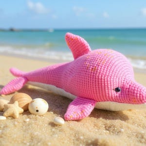 May include: A pink crochet dolphin toy with a white underside, resting on a sandy beach with seashells. The dolphin has black eyes and yellow flecks. The sea and sky are visible in the background.
