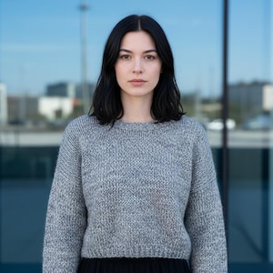 May include: A woman with dark hair wearing a gray knit sweater. The sweater has a crew neck and long sleeves. The background is a modern building with large glass windows.