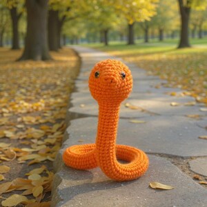 Peut inclure: Un jouet serpent orange crocheté avec des yeux noirs. Le serpent est fait de fil et a un corps long et mince avec une tête arrondie. Il est posé sur une surface en pierre. L'arrière-plan montre un parc flou avec un chemin et des arbres.