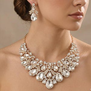 May include: A gold-toned statement necklace and matching drop earrings featuring clear, pear-shaped and round faceted crystals. The necklace has a wide, layered design with multiple crystal clusters, while the earrings mirror the top section.