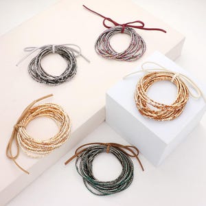 May include: Five circular hair ties featuring multiple strands of sparkling rhinestones in silver, gold, dark green, and multicolored tones, each secured with a thin, coordinating suede-like cord tied into a small bow at the top.