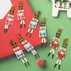 May include: A collection of dangling nutcracker earrings featuring articulated limbs and gold glitter star toppers. The nutcrackers are painted in various colors including pink, light blue, and red, each wearing a black hat and uniform details.