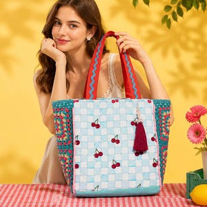 May include: A woven tote bag with a light blue and white checkered pattern, featuring embroidered cherries. The bag has red handles with blue trim and a red tassel. The sides are crocheted with a floral pattern.