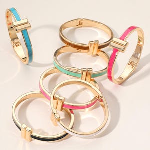 May include: A collection of gold-tone metal bangles with colorful enamel accents. The bracelets feature a rectangular clasp and come in various colors, including turquoise, brown, mint green, pink, black, and white. The bangles are arranged on a light-colored surface.
