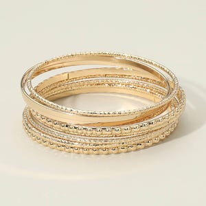 May include: A stack of gold-colored bangles. The bangles have varying textures, including smooth, ridged, and dotted patterns. The jewelry is arranged in a circular shape, with multiple bangles visible.