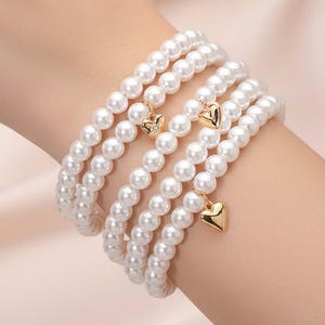 May include: Four white pearl bracelets are stacked on a wrist. Each bracelet is strung with round, white pearls. Gold heart charms dangle from three of the bracelets. One charm has a small diamond. The bracelets are a jewelry accessory.