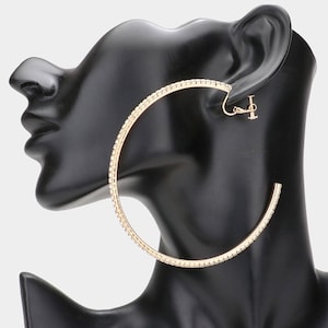 May include: Gold-toned hoop earrings with a row of small, clear crystals. The earrings are large and circular, with a clip-on closure. The earrings are displayed on a black mannequin head.
