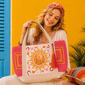 May include: A pink tote bag with a sun design. The bag has a beige canvas base and handles, with a crochet pink border. The sun design is in the center, with orange and yellow accents. A tassel hangs from the side.