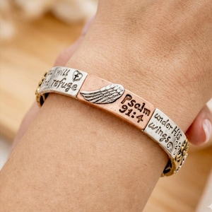 May include: A silver, copper, and gold-toned bracelet with religious inscriptions. The bracelet features the text "You will find refuge", a wing design, and "Psalm 91:4 under His wings". The bracelet is worn on a wrist.