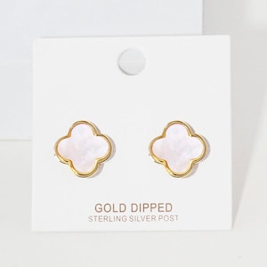 May include: A pair of stud earrings featuring a four-leaf clover shape with a white iridescent inlay and a gold-toned metal border. The earrings are attached to a white display card with the text GOLD DIPPED STERLING SILVER POST printed below.