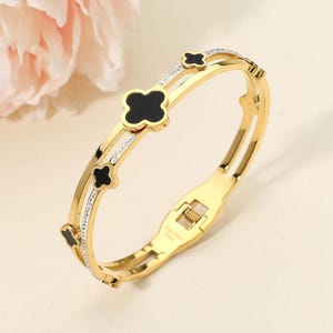 May include: A gold-toned hinged bangle bracelet featuring a double-band design. The bracelet is accented with four black clover-shaped motifs and a single row of small clear crystals embedded along the inner band, secured with a metal clasp.