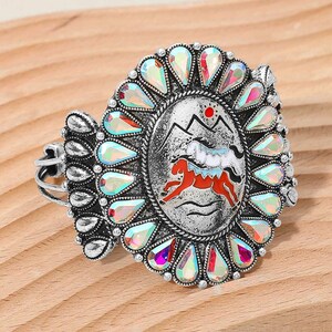 May include: Silver-toned cuff bracelet with an oval centerpiece featuring a mountain scene and two horses. The bracelet is adorned with iridescent teardrop-shaped stones around the oval. The horses are red and white.