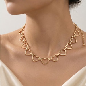May include: Gold-colored necklace featuring a series of open heart-shaped links. The necklace is worn around the neck, with the hearts evenly spaced. The chain has an adjustable length and a small ball at the end. The jewelry is a simple yet elegant accessory.