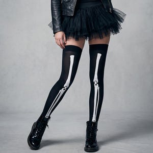 May include: Black thigh-high stockings featuring a white printed skeleton leg bone design that runs vertically down the front of each leg. The stockings are paired with a black tulle skirt, a black leather jacket, and black lace-up boots.