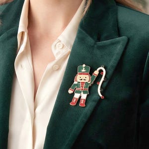 May include: A nutcracker brooch featuring a green and white uniform with red accents, a black mustache, and a curved white staff adorned with small red crystals. The enamel pin is attached to the lapel of a dark green velvet blazer over a white shirt.