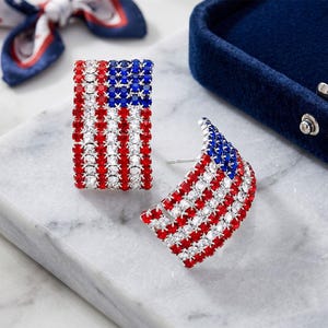 May include: A pair of curved hoop earrings featuring a pattern of red, white, and blue rhinestones arranged to resemble the American flag. The earrings have a post-back closure and are designed with a semi-circular shape and a sparkling finish.