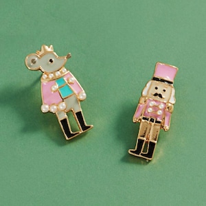 May include: Two gold-tone metal enamel pins. One pin features a mouse wearing a pink and teal jacket with white pearl-like accents. The second pin depicts a nutcracker figure in a pink uniform with a tall hat and white pearl-like buttons.