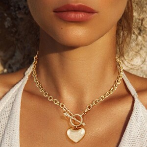 May include: A gold-colored necklace with a heart-shaped pendant. The necklace features a toggle clasp and a chain link design. The heart pendant is smooth and shiny, and the necklace is worn around the neck.
