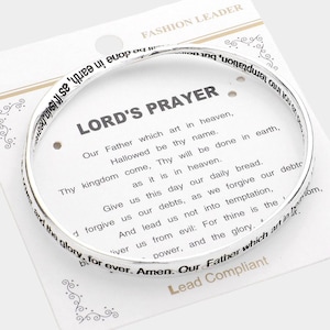 May include: A silver-colored bangle bracelet with the Lord's Prayer inscribed around the band. The bracelet is displayed on a white card with the prayer printed in black text. The card also has the words "Fashion Leader" at the top.