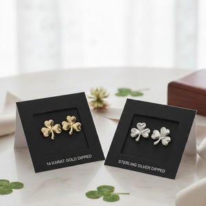 May include: Two pairs of clover-shaped stud earrings, one gold-dipped and the other sterling silver, displayed on black cards. The gold earrings are labeled "14 Karat Gold Dipped," and the silver earrings are labeled "Sterling Silver Dipped."