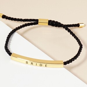 May include: A black braided bracelet with a gold-colored rectangular centerpiece. The word "BRIDE" is engraved on the centerpiece. The bracelet has gold-colored metal accents at the ends and adjustable sliding knots.