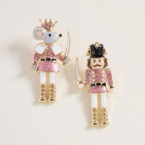 May include: Two gold-tone metal pins featuring a mouse king and a nutcracker soldier. Both figures wear pink glitter uniforms with white accents, gold detailing, and carry small swords. The mouse wears a gold crown, while the soldier has a black hat.