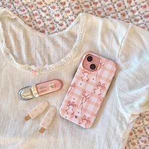 Op de afbeelding: Een witte, getextureerde top met korte mouwen en een kanten halslijn, gecombineerd met een roze geruite telefoonhoes met strikjes. Bevat ook een roze lipgloss en twee ijslolly-vormige haarclips.