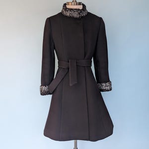 May include: A black coat with a flared skirt, long sleeves, and a matching belt. The coat features a faux fur collar and cuffs. The coat is displayed on a vintage dress form against a light blue background.