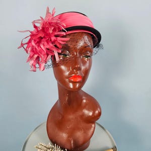 Puede incluir: Un sombrero rosa vintage con un borde negro y un acento decorativo de plumas rosas. El sombrero presenta un velo de red negro y se exhibe en una cabeza de maniquí marrón. Un ramo de flores secas está en primer plano.