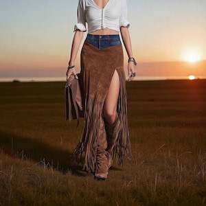 May include: A brown suede skirt with a denim waistband and a thigh-high slit, featuring fringe detailing. The skirt is worn with brown cowboy boots and a white top. A brown leather bag is held. The background is a field at sunset.
