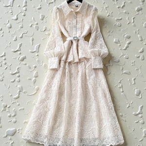 Könnte beinhalten: Ein cremefarbenes Spitzenkleid mit langen, durchsichtigen, gepufften Ärmeln und einem Kragen. Das Kleid hat ein figurbetontes Oberteil, einen weißen Gürtel mit dekorativer Schnalle und einen fließenden Rock. Das Kleid hängt an einem schwarzen Kleiderbügel vor einer strukturierten Wand.