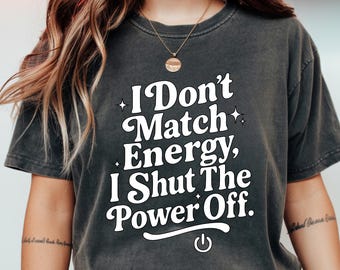May include: Dark gray t-shirt with the text "I Don't Match Energy, I Shut The Power Off." in white. The shirt has a crew neck and short sleeves. The text is in a bold, stylized font with a power button graphic.