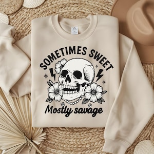 Op de afbeelding: Beige sweatshirt met een afbeelding van een schedel, bloemen en bliksemschichten. De tekst luidt "Sometimes Sweet, Mostly Savage". Het ontwerp is in zwart en wit. De sweatshirt ligt op een geweven oppervlak.