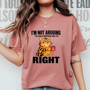 Puede incluir: Una camiseta color rosa empolvado con el gráfico de un gato atigrado naranja que sostiene una taza roja con el texto "COFFEE MY RULES". Sobre el gato, el texto dice "I'M NOT ARGUING I'm just explaining why I'm" y debajo, la palabra "RIGHT".