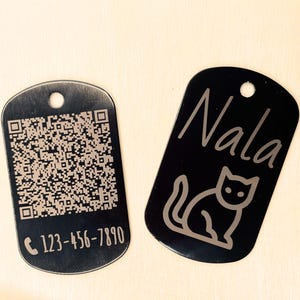 Peut inclure: Deux plaques d'identification noires pour animaux de compagnie. L'une comporte un code QR et un numéro de téléphone. L'autre porte le nom "Nala" et une silhouette de chat. Les deux ont un trou en haut.
