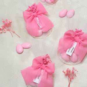 Pode incluir: Sacos de lembrancinhas de tule rosa com amêndoas açucaradas rosa, amarrados com laços rosa e adornados com flores secas. Cada saco tem uma pequena etiqueta com texto. As lembrancinhas são dispostas em uma superfície branca.