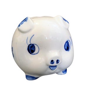 May include: A white ceramic piggy bank with blue accents. The pig has blue eyes, ears, and small details. The piggy bank is round with a smiling face and small feet.