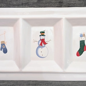 May include: White rectangular serving dish with three square compartments. Each compartment features a Christmas-themed illustration: blue mittens, a snowman with a red scarf, and a green stocking with gifts.